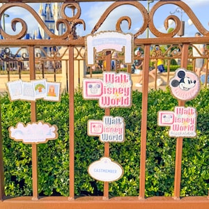 May include: A collection of Disney-themed patches and signs, including "Walt Disney World" and a Mickey Mouse design. The items are displayed on a brown metal fence with a castle in the background. The signs are in pink, white, and gold.