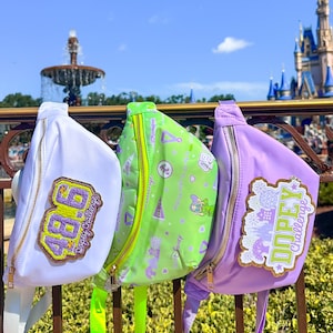 May include: Three colorful fanny packs with gold zippers and patches. The fanny packs are white, green, and purple. The white fanny pack has a patch that says "48.6 Dopey Challenge". The green fanny pack has a pattern and the purple fanny pack says "Dopey Challenge".