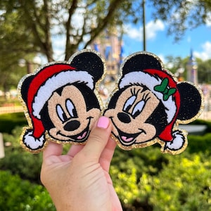 May include: Two embroidered patches featuring Mickey and Minnie Mouse wearing Santa hats. The patches have black ears, tan faces, and red and white Santa hats. A gold glitter border surrounds each patch. The background shows a blurred outdoor scene.