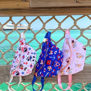 Puede incluir: Tres riñoneras con cremalleras doradas y correas ajustables cuelgan de una barandilla de madera. Las riñoneras presentan diseños náuticos y de personajes de dibujos animados en rojo, azul y rosa. El fondo muestra un crucero y el mar.