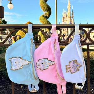May include: Three fanny packs with different colored fabric and a gold zipper. The fanny packs have a sparkly gold castle design with a pink bow on the white fanny pack.