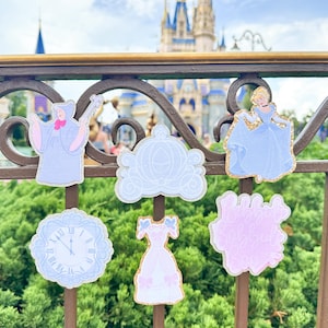 May include: A collection of Disney-themed decorative pins featuring Cinderella and related imagery. The pins include Cinderella's dress, carriage, and a clock, all in pastel colors with glitter accents. The background shows a castle and lush greenery.