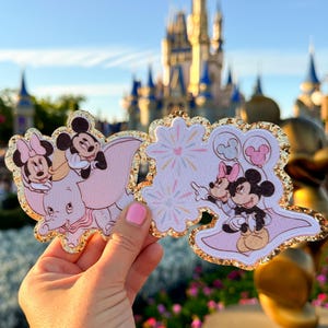 以下が含まれることがあります： ミッキーマウスとミニーマウスをフィーチャーしたピンクとゴールドのグリッターアイロン接着パッチ2枚。最初のパッチは、ミッキーとミニーがダンボに乗っている様子を描いています。2枚目のパッチは、ミッキーとミニーが風船を持ち、背景に花火が描かれています。
