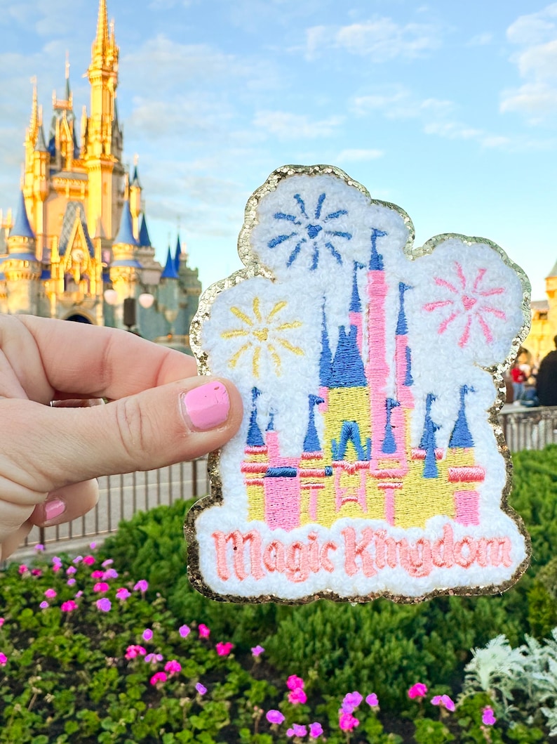 May include: A white felt patch with gold glitter trim featuring an embroidered image of Cinderella's Castle with fireworks and the text "Magic Kingdom".