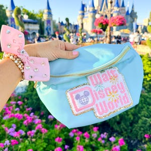 May include: A light blue fanny pack with a pink and gold "Walt Disney World" patch featuring a Mickey Mouse head with a globe. The fanny pack is held in front of a blurred background of Cinderella's Castle at Walt Disney World.
