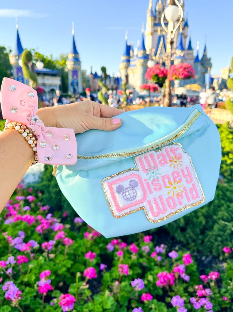 May include: A light blue fanny pack with a pink and gold "Walt Disney World" patch featuring a Mickey Mouse head with a globe. The fanny pack is held in front of a blurred background of Cinderella's Castle at Walt Disney World.