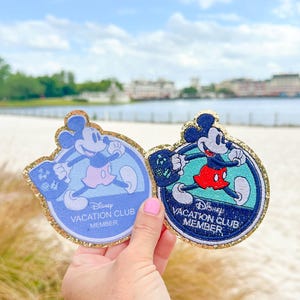 May include: Two round Disney Vacation Club Member patches featuring Mickey Mouse. The patches are blue and green with gold glitter trim. The text on the patches reads "Disney Vacation Club Member".
