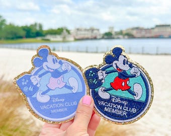 Può includere: Due toppe rotonde Disney Vacation Club Member con Mickey Mouse. Le toppe sono blu e verdi con un bordo di glitter dorato. Il testo sulle toppe recita "Disney Vacation Club Member".