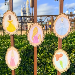 May include: Six decorative oval-shaped ornaments featuring Disney princesses. Each ornament has a gold-colored scalloped border and a light blue or pink background. The princesses are depicted in their iconic dresses, with a castle in the background.