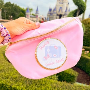 May include: A light pink fanny pack with a gold zipper and a circular patch featuring a blue suitcase, a pair of mouse ears, and the text Disney Travel Agent. The patch is bordered by a gold glitter trim.