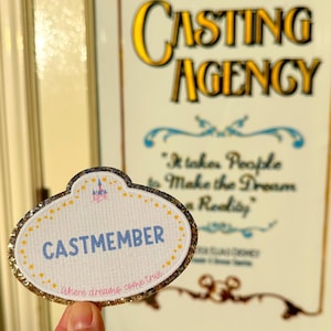 May include: A Cast Member name tag with a gold glitter border and the word "CASTMEMBER" in blue. The tag is held in front of a sign that reads "Magic Kingdom CASTING AGENCY". The phrase "Where dreams come true" is also visible.