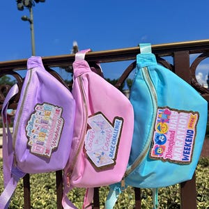 May include: Three colorful fanny packs in purple, pink, and blue, each with a gold zipper and a matching strap. The packs feature embroidered patches with text and graphics, including "Disneyland Weekend" and "Coast to Coast Race Challenge."