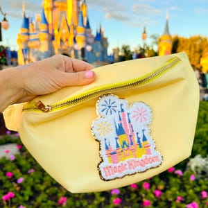 May include: A yellow fanny pack with a sparkly pink, yellow, and blue embroidered patch of Cinderella's castle with fireworks and the text "Magic Kingdom".