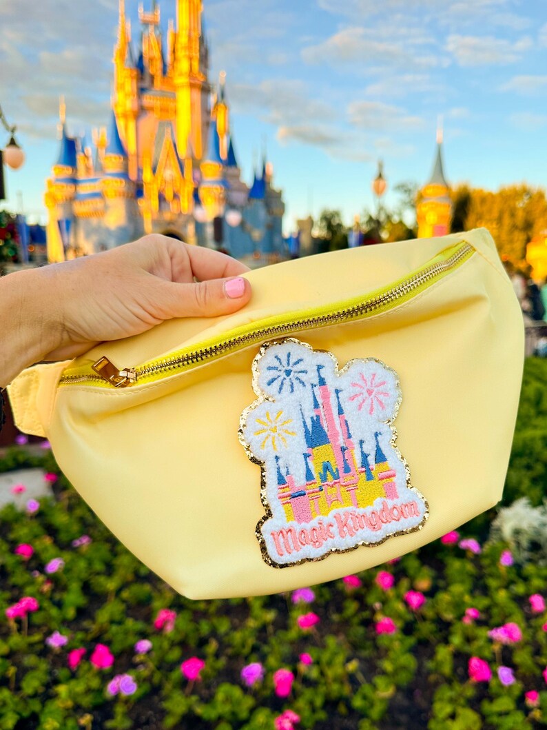 May include: A yellow fanny pack with a sparkly pink, yellow, and blue embroidered patch of Cinderella's castle with fireworks and the text "Magic Kingdom".