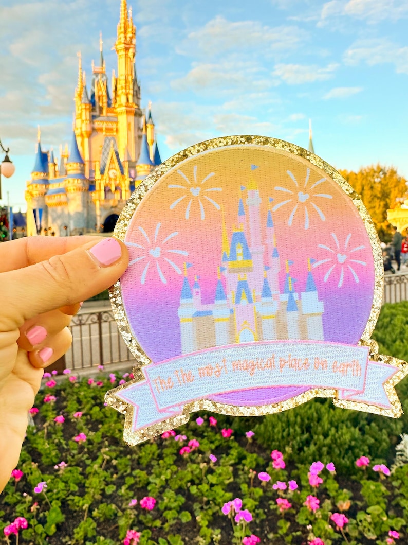 May include: A pink and purple glitter iron-on patch with a silhouette of Cinderella's castle and fireworks. The text "the most magical place on earth" is on a banner below the castle.