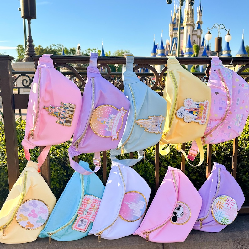 May include: A collection of eight pastel-colored fanny packs with various Disney-themed patches. The patches feature images of Cinderella's castle, Mickey Mouse, fireworks, and other Disney icons.