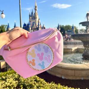 May include: A pink fanny pack with a gold zipper and a round, white patch featuring colorful Mickey Mouse balloons. The patch has a gold glitter border.