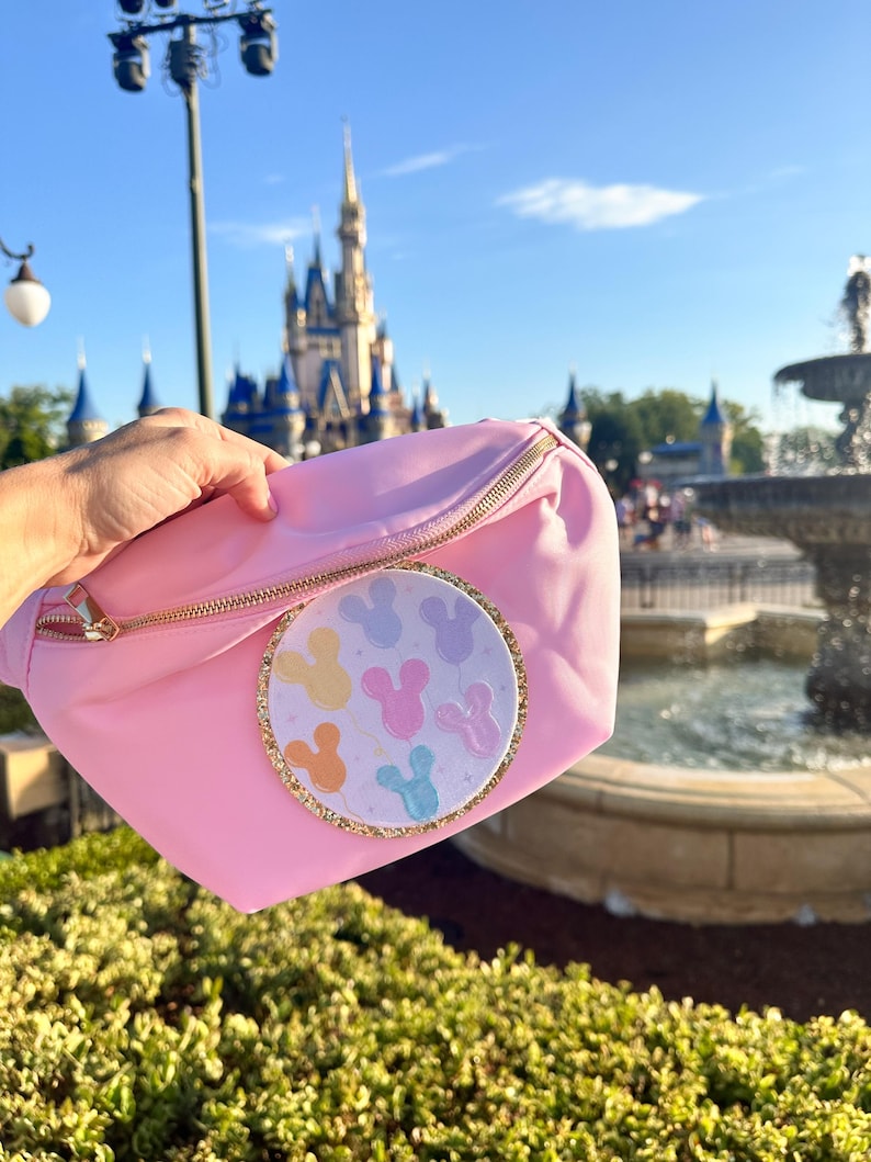 May include: A pink fanny pack with a gold zipper and a round, white patch featuring colorful Mickey Mouse balloons. The patch has a gold glitter border.
