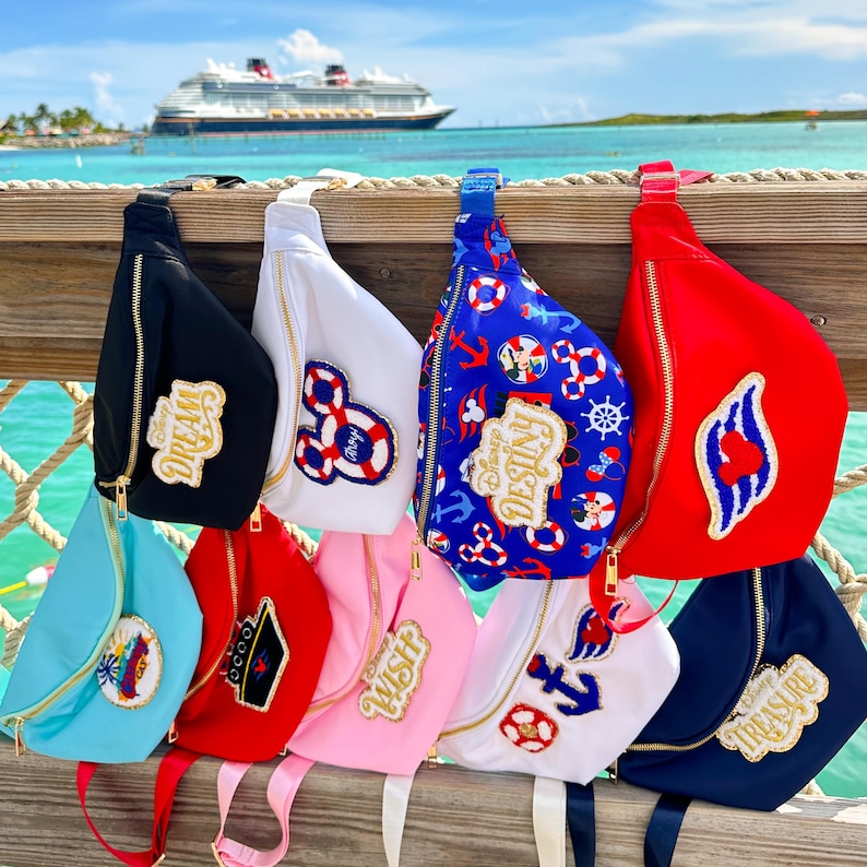 May include: A collection of ten belt bags in various colors including black, white, blue, red, light blue, and pink. Each bag features a gold zipper and a unique embroidered patch with designs like anchors, life preservers, and text like Dream, Destiny, Wish, and Treasure.