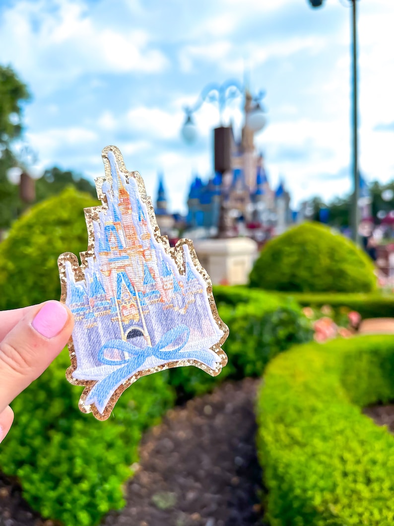 May include: A sparkly gold glitter iron-on patch featuring a detailed illustration of Cinderella's castle with a blue bow.