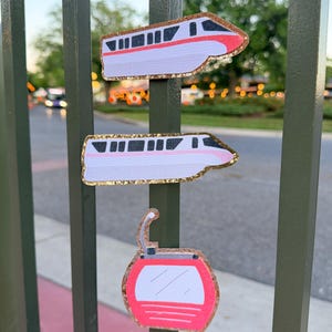 May include: Three decorative stickers: two monorail train designs and a red and white gondola car. The stickers have gold glitter edging and are affixed to a green metal fence. The background is blurred, suggesting an outdoor setting.