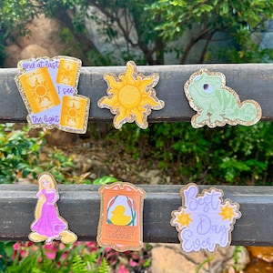 Op de afbeelding: Een verzameling kleurrijke, glinsterende patches met personages en symbolen uit een populaire animatiefilm. Ontwerpen omvatten een zon, een kameleon, een prinses in een roze jurk, lantaarns met de tekst "and at last I see the light", een bord voor "The Snuggly Duckling" en de zin "Best Day Ever".