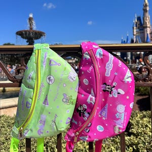 May include: Two fanny packs, one lime green and one hot pink, with gold zippers and straps. The green pack features Disney-themed graphics and text, while the pink pack showcases Minnie Mouse and other Disney icons. Both are hanging on a metal railing.