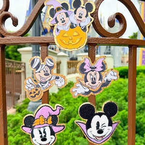 May include: Five embroidered patches featuring Disney characters in Halloween costumes. The designs include Mickey and Minnie in a pumpkin, a mummy Mickey, a vampire Minnie, a witch Minnie, and a vampire Mickey, all with gold glitter borders.