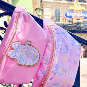 May include: Two pink fanny packs featuring Ratatouille-themed designs. One bag is solid pink with a glittery patch of Remy in a chef hat. The other bag has a repeating pattern of Remy, the Eiffel Tower, and the text "anyone can cook" and "Gusteau's".