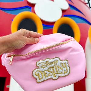 May include: A light pink fanny pack featuring a gold zipper and a textured patch with white and gold glitter embroidery that reads Disney Destiny. The bag has a smooth, matte finish and a single main compartment with a gold metal zipper pull.