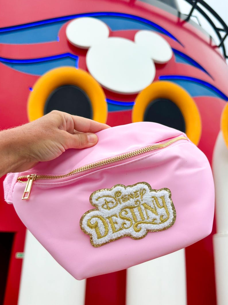 May include: A light pink fanny pack featuring a gold zipper and a textured patch with white and gold glitter embroidery that reads Disney Destiny. The bag has a smooth, matte finish and a single main compartment with a gold metal zipper pull.