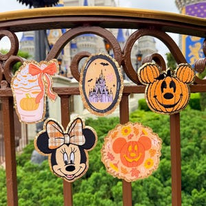 May include: Five decorative patches featuring autumn and character themes. Designs include a pumpkin-topped drink, a castle in an oval frame, a pumpkin-shaped character head, a mouse character with a plaid bow, and a pumpkin surrounded by fall leaves.