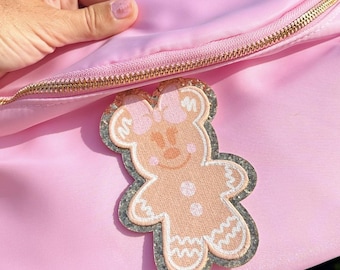 Puede incluir: Una riñonera de color rosa claro con una cremallera dorada y un parche central con forma de personaje de pan de jengibre con un lazo rosa, detalles de glaseado blanco y un borde de purpurina plateada, hecha de material sintético liso.
