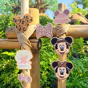 May include: A collection of embroidered patches featuring a leopard print Mickey Mouse head, a leopard print bow, a safari hat, a tree with the text ANIMAL KINGDOM, a small pair of ears, and two character faces wearing safari hats with ears.