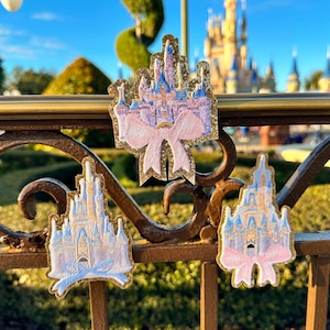 Könnte beinhalten: Drei gestickte Aufnäher mit einem rosa und blauen Schloss mit einer rosa Schleife. Die Aufnäher befinden sich auf einem braunen Metallgeländer mit einem verschwommenen Hintergrund eines Schlosses.