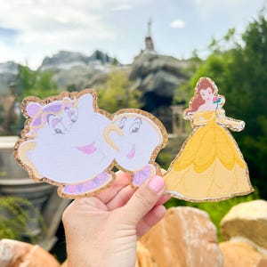 Könnte beinhalten: Ausgeschnittene Figuren der Disney-Figuren aus Die Schöne und das Biest, Mrs. Potts und Chip, sowie Belle in ihrem gelben Ballkleid. Die Figuren haben glitzernde Goldränder und werden vor einem verschwommenen Hintergrund im Freien gehalten.