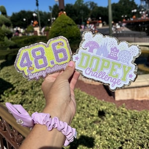 May include: Two embroidered patches with glitter borders. One patch displays the number "48.6" in purple on a lime green background, with "Dopey Challenge" text. The other patch features a castle and fireworks with "Dopey Challenge" text. A lavender scrunchie is also visible.
