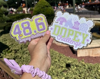Könnte beinhalten: Zwei bestickte Aufnäher mit goldglitzernden Rändern. Ein Aufnäher zeigt den violetten Text 48.6 und Dopey Challenge. Der andere Aufnäher zeigt den hellgrünen Text Dopey Challenge mit violetten Silhouetten von Park-Symbolen und Feuerwerk.