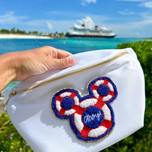 May include: A white belt bag featuring a gold zipper and a large, embroidered patch shaped like a mouse head. The patch is designed as a red, white, and blue life preserver with the word Ahoy! stitched in blue thread in the center.