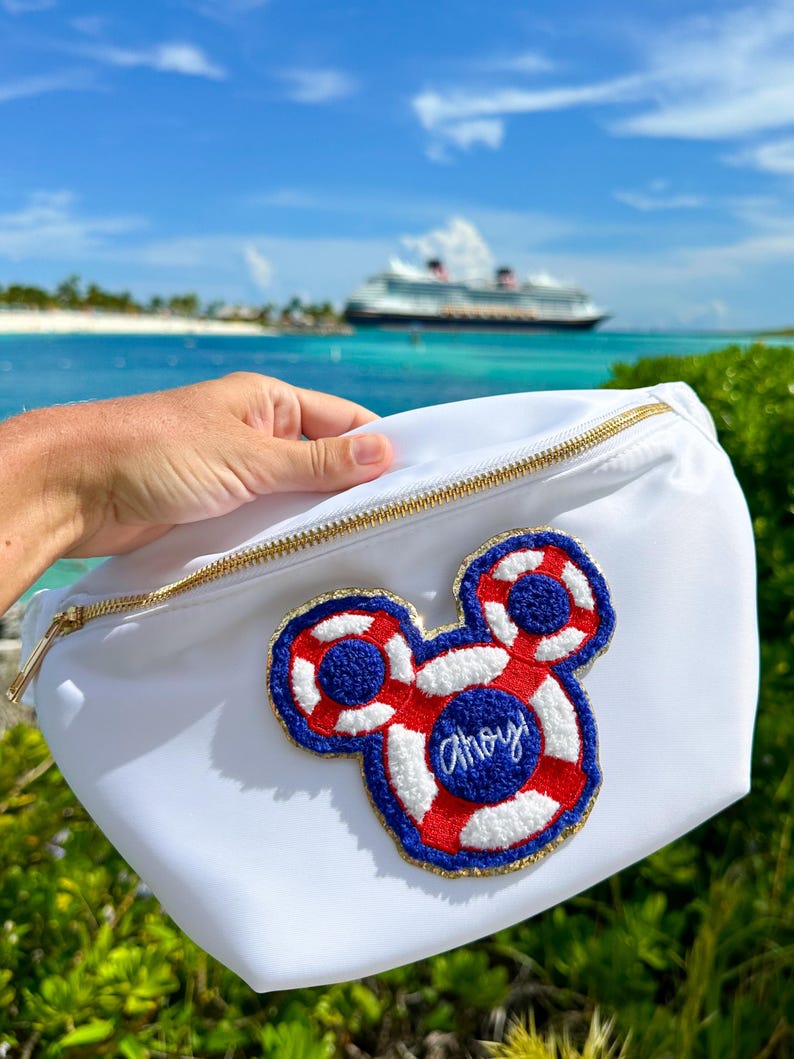 May include: A white belt bag featuring a gold zipper and a large, embroidered patch shaped like a mouse head. The patch is designed as a red, white, and blue life preserver with the word Ahoy! stitched in blue thread in the center.