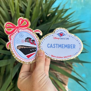 May include: A Disney Cruise Line Cast Member name tag and a matching oval ornament featuring a cruise ship illustration. The name tag is white with blue text reading CASTMEMBER and Where dreams come true, accented by gold glitter borders.