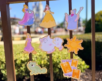 May include: A collection of eight die-cut stickers featuring animated characters and symbols, including a princess in a pink dress, a princess in a yellow dress, a fairy godmother, a long-haired princess, a teapot, a sun, a lizard, and lanterns with text.