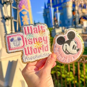 Könnte beinhalten: Zwei gestickte Aufnäher mit goldfarbenem Rand. Ein Aufnäher trägt die Aufschrift "Walt Disney World Passholder" mit einem Mickey Mouse-Symbol. Der andere Aufnäher zeigt ein rosa Mickey Mouse-Gesicht. Beide Aufnäher werden vor einem Schloss gehalten.