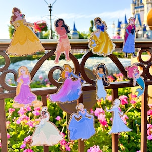 Może przedstawiać: Kolekcja 11 naklejek z postaciami księżniczek Disneya, w tym Belle, Śnieżka, Ariel i Mulan. Naklejki znajdują się na metalowym ogrodzeniu z rozmytym tłem parku.