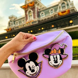 May include: A light purple fanny pack featuring two embroidered patches: one of Mickey Mouse dressed as a vampire with a purple cape, and one of Minnie Mouse wearing a purple witch hat with an orange bow, both outlined in gold glitter thread.
