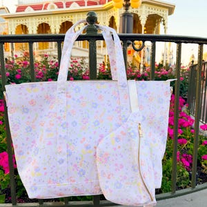 May include: A white tote bag and a matching fanny pack, both featuring a repeating pastel floral pattern in shades of light pink, yellow, and blue. The tote has two top handles and a zipper closure, while the fanny pack has a gold-tone zipper.