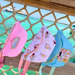 May include: Three fanny packs hanging on a rope railing. The left bag is solid pink with a cruise ship patch. The center bag is white with a repeating pattern of cartoon characters. The right bag is light blue with a patch featuring multiple characters.