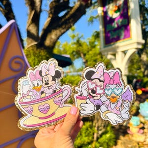 Puede incluir: Dos parches brillantes con Minnie Mouse y Daisy Duck. Uno los muestra en una taza de té, el otro con gafas de sol y bebidas. Los parches tienen detalles en rosa, amarillo y dorado, perfectos para los fans de Disney.