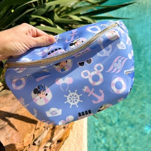 May include: A light blue fanny pack with a gold zipper, featuring a nautical-themed pattern with anchors, Mickey Mouse, and life preservers. The bag is held by a hand, with a pool in the background.