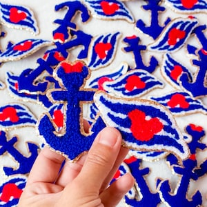 May include: A collection of embroidered patches featuring a blue anchor with a red bow and a blue and white striped wing with a red heart shape. Each patch is outlined with a gold metallic border, creating a textured and decorative appearance.